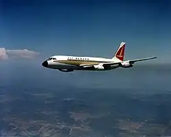 Photo couleur d'un avion en vol vu de trois quart face. Le ciel est presque totalement dégagé et un relief vallonné se situe sous l'appareil, qui se trouve à haute altitude.
