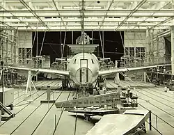 Cliché noir et blanc. Hangar avec de très nombreux éléments d'assemblage. Au centre avion en cours de montage. Pas de nez.