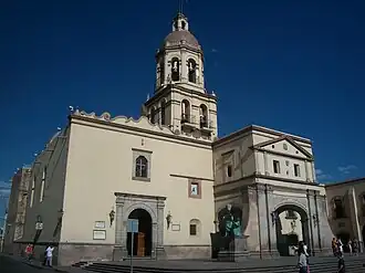 édifice mêlant les styles néoclassique et baroque de couleur claire devant un ciel azur