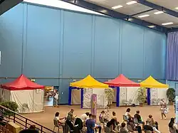 Quatre barnums alignés contre un mur dans le vaste hall d'un gymnase devant lesquels attendent des personnes en attente de vaccination.