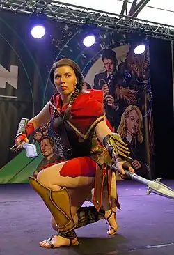 Photo d'une femme déguisée en Kassandra, portant une tenue de mercenaire rouge et dorée, la lame de Léonidas et une épée, et étant agenouillée.