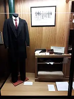 Photo d'une vitrine où est exposé un costume masculin de la fin du XIXe&nbsp;siècle. Il est formé d'un ensemble en drap de laine noir comprenant une redingote, un gilet et un pantalon, une chemise blanche ornée en coton d'un plastron, d'un col amovible et d'une cravate nouée en soie. Sur la droite, un support bas expose des accessoires tels qu'un coffret à boutons de manchettes et des gants blancs dans leur boîte.