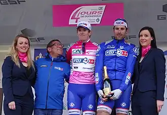 Trois hommes habillés de bleu et de rose, entourés de deux personnes, sourient, en tenant une bouteille de champagne.