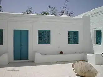Cour intérieure d'un houch avec ses murs blancs, sa porte et ses grilles aux fenêtres de couleur bleue.