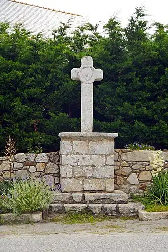 Vue d’une croix de chemin, avec pied en pierres de taille sur fond de haie de thuyas.