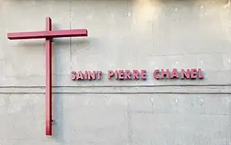 Croix rouge et inscription "Saint Pierre Chanel" en rouge, sur un mur en béton.