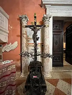 Photographie couleur d'un crucifix en ferronnerie. En arrière-plan, la porte d'entrée en bois massif et deux colonnes en marbre.