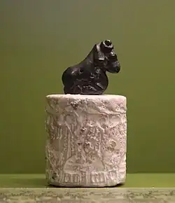 Cylindre gravé de couleur claire, surmonté par une figurine d'un bélier accroupi.