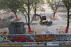 Au premier plan, le mur de 2,5 mètres de hauteur s'étend sur toute a longueur de la photo. On y voit un certain nombre de graffitis, l'un d'entre-eux dénonce le coût de construction du mur, soit 390 000 €. Un autre affirme "CHANTIER SOLEAM QUARTIER EN DANGER" avec une petite tête de mort, vestiges visibles de la contestation du chantier qui commence sur la place. Au second plan, un engin de chantier désolidarise le revêtement de pavés qui tapissait jusqu'alors le sol de la place, révélant un sol terreux. À l'arrière plan, on distingue la partie opposée du mur de l'autre côté de la place.