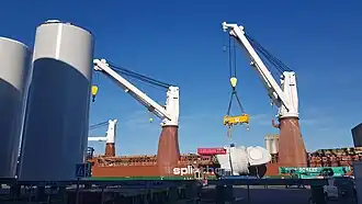 À l'arrière plan, la grue du navire de transport vient de déposer la turbine sur son ber. La grue est encore équipée du palonnier. Au premier plan de gauche à droite on voit des sections des piles des futurs éoliennes, et des bers vide pour en accueillir de nouveaux. Au premier plan au milieu à gauche on distingue une plateforme automotrice (verte) qui sert à déplacer les éléments, et à droite à l'arrière plan une des grues mobiles (verte) qui permet de manipuler les éléments... Cette grue mobile n'est pas suffisante pour déplacer les piles ou les turbines, une grande structure porteuse (non photographiée ici) sert à redresser les piles, qui arrivent horizontales par bateau, et sont redressées avant d'être stockées verticales.