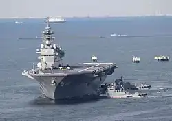 Photo couleur d'un porte-avions naviguant sur une mer calme, flanqué, à sa droite, de deux navires plus petits.