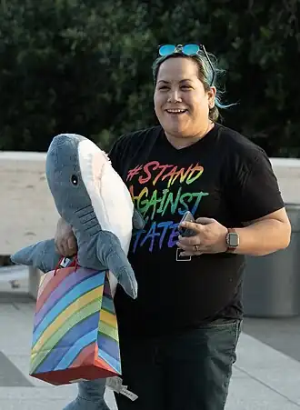 Photographie d'une femme aynt des cheveux longs attachés en chignon dont une partie sont teints en bleus, un tshirt noir avec "Stand against hate" écrit en arc-en-ciel et un jean. Elle tient contre elle un requin en peluche et tient dans la main un sac en papier arc-en-ciel