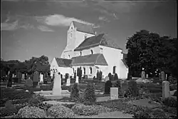 Photo en noir et blanc d'église ancienne, éclairée par une lumière douce au cœur d'un cimetière verdoyant.