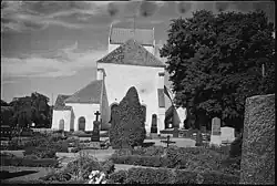 Photo en noir et blanc. Église blanche , parmi tombes et fleurs, dans une ambiance mélancolique.