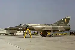 Un Mirage 5 immobile au sol, sur un tarmac, avec d'autres avions stationnés à l'arrière-plan.