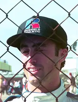 Photo d'un homme souriant, portant une casquette noire, derrière un grillage