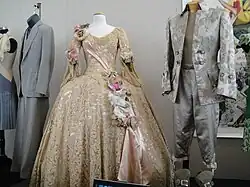 Une robe beige très ornementée, entre autres de grosses fleurs, et un costume gris masculin en satin lui aussi décoré d'un motif floral imprimé. Les deux sont exposés dans une galerie.