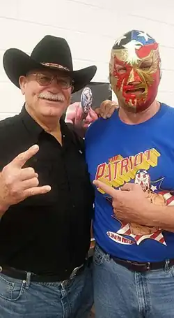 Un homme moustachu portant un stetson et une chemise noire et un autre masqué portant un t-shirt bleu.