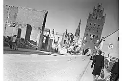 Photo en noir et blanc prise de travers, montrant des femmes marchant sur un trottoir à gauche, et des ruines de maisons brûlées, à droite.