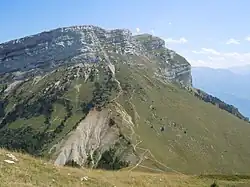 Barres rocheuses dominant une grande pente herbeuse parcourue d'un sentier en zigzag.