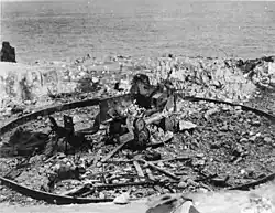 Photographie en noir et blanc de restes en métal calciné et en petits morceaux. Une structure métallique en cercle est visible.