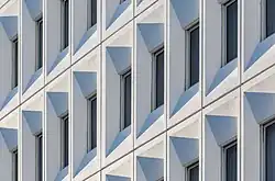 Des rangées de fenêtres toutes pareilles sur un mur blanc forment une géométrie de gris.