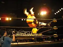 Un catcheur effectue une prise du haut de la troisième corde en portant son adversaire sur ses épaules. Ils sont en l'air et s'apprêtent à passer à travers un panneau de bois recouvert de fils barbelés.