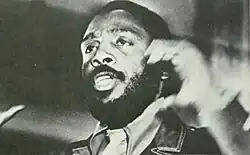 Vue en légère contre-plongée d'un homme barbu de trois-quart agitant une main, cadré au niveau des épaules