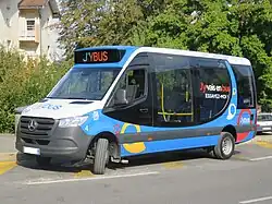 Photographie en couleurs d’un minibus bleu, blanc et noir de type Dietrich City 29, marqué J'ybus, et stationné à proximité de bâtiments résidentiels