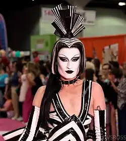 Drag queen en robe blanche et noire, avec du masacara blanc et du rouge à lèvre noir.