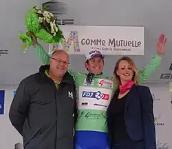 Trois personnes se tiennent alignées. Celle du centre, habillée en vert pomme, lève les bras avec un bouquet.