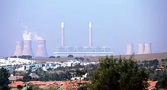 Au premier plan des maisons basses ; au fond, derrière des collines pelées, une usine avec 2 grandes cheminées et 6 tours de refroidissement.