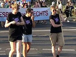 Au premier plan, trois lesbiennes portant des cheveux courts, un tshirt noir avec "Dyke" écrit sur une poing violet. L'une porte un short noir, l'autre un short en jean et la troisième un cargo short kaki. Au second plan, une dizaine de lesbiennes portent une banderole. Elles ont toutes les cheveux courts.