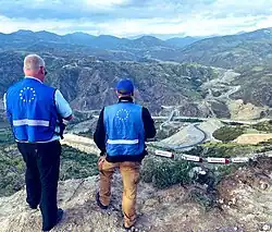 Photographie de deux hommes au bord d'une falaise, observant en contre-bas des camions arrêtés sur une route de montagne.