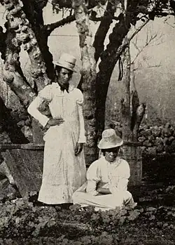 Photographie en noir et blanc de deux femmes. Une femme se tient debout, l'autre est assise. Elles portent une robe blanche et un chapeau typiquement victorien.