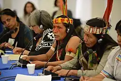 Cinq femmes sont installées à une tribune officielle équipée de micros. Parmi elles, deux portent des ornements remarquables comprenant des plumes et d'autres accessoires. On distingue d'autres personnes à l'arrière-plan et à l'avant-plan.