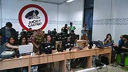 Une foule de personnes assises devant des ordinateurs travaillent dans une salle aveugle. Sur le mur, en arrière plan est affiché le slogan « Procès et sanction» en espagnol.