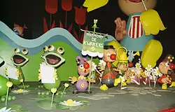 Scène intérieure composée de personnages fictifs. L’ensemble a un aspect enfantin, digne d’un dessin animé. Ceux-ci ne possèdent ni bras, ni jambes, à la manière des dessins d’enfants. Sur la gauche, trois grenouilles humanoïdes tiennent une partition en mains et semblent la chanter, leur bouche étant grandes ouvertes. À leurs pieds sont posés des nénuphars artificiels. Ensuite, à droite se trouvent trois personnages masculins. Tous les personnages représentant des êtres humains portent un nez de clown et des petites dalles de pierre sont posées à leur pied. Le premier est le plus petit. Habillé des pieds à la tête de violet, les exceptions sont ses souliers noirs, un bouton jaune sur le ventre, une collerette jaune à clochettes, des gants jaunes et deux cloches aux extrémités de son bonnet pointu. Il tient dans sa main droite un étendard dont l’écriteau imite un tissu vert à franges blanches. Sur celui-ci est inscrit Harmony de kwakkers. Le second représente un fermier avec son gilet marron foncé, sa chemise rouge, son pantalon à fines rayures verticales blanches et bleues foncées, ses sabots jaunes et sa casquette noire. Le troisième personnage est habillé de noir. Il porte également des sabots jaunes mais aussi une collerette verte, des cheveux verts, et en guise de chapeau, un arrosoir jaune. Quelques fleurs artificielles le séparent du quatrième personnage fumant la pipe, à l’extrême droite. Il porte aussi les sabots jaunes, mais son pantalon est noir, sa chemise rouge et son chapeau bordeaux. Trois boutons disposés en triangle sont sur la chemise et sur le chapeau. Au second plan, un panneau vert forme le fond. Son bord supérieur est découpé en forme de vague. Deux rayures, bleu clair et bleu foncé, la suivent. Trois grandes tulipes rouges sont situées derrière ce panneau. Un personnage aux proportions bien plus grandes occupe le fond de la photo. L’observateur ne le voit que jusqu'à son menton sur la photo. Habillé d’un pantalon noir, d’une chemise bleue avec bavoir rayé verticalement de blanc et rouge et de sabots jaunes, deux tulipes sont situées à 5H et à 8H par rapport à son visage.