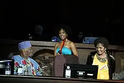 Une jeune-femme est debout, avec une robe bleu clair, entre deux personnes plus âgées, un homme à gauche assis, vêtu de tissu coloré et portant un couvre-chef  bleu à motifs géométriques, et une femme à droite debout, portant de façon visible une décoration aux couleurs du drapeau de l'Équateur