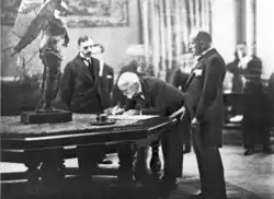 Un homme chauve penché sur une table de marbre signe un document tandis que deux hommes l'observent.