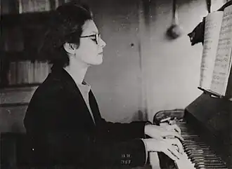 Photographie en noir et blanc d'une femme de profil, portant des lunettes et un smoking, au piano.