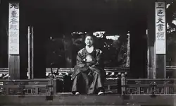 Photographie en noir et blanc montrant un homme assis sous un auvent en bois et vêtu d’une robe richement brodée de dragons sur la poitrine et les épaules.