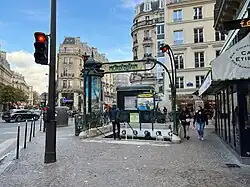 Photographie en couleurs d'une bouche de métro, avec portique en fer frogé très large et perspective diagonale d'une rue se prolongeant vers le fond de l'image