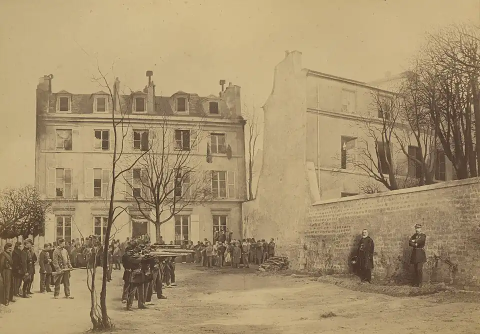 Photographie montrant deux hommes debout devant un mur face à des soldats qui mettent en joue leurs fusils.