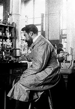 Photographie d’un homme brun, portant barbe et moustache, vu du côté gauche, assis sur un tabouret haut, devant une paillasse de laboratoire.