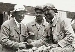 Photo de trois hommes, l'un en tenue de pilote reçoit les félicitations d'un autre en costume, sous le regard d'un mécanicien