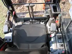 Poste de conduite d'une pelleteuse sur chenilles : manettes-pédales séparées d'avancement des chenilles au centre, joysticks de commande de la tourelle et de la flèche, et de l'outil, sur les côtés.
