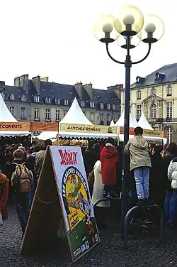 Photographie d'une place de Rennes pendant la "Fête d'Astérix et Obélix". De nombreuses personnes ainsi que des tentes pour les stands et un panneau "Astérix" sont visibles. Notons, sur le panneau, l'inscription "Les marchés de Condate".