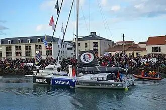 Bateau noir, bleu et gris. Le navigateur, de dos, salue des deux bras la foule massée sur le quai.