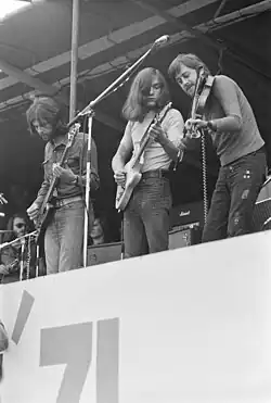 Photo en noir et blanc et en contreplongée de trois musiciens au bord d'une scène : un bassiste, un guitariste et un violoniste. On distingue à l'arrière-plan des roadies et des amplis.